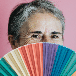 An older woman happily peers over an open rainbow hand fan.