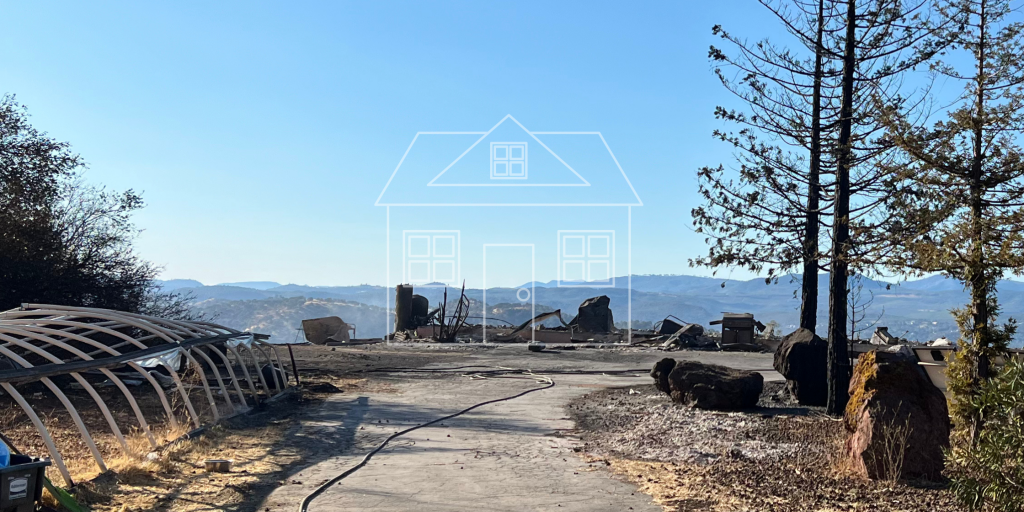 Image depicting a ghostly graphic outline of a house overlayed on top of the remains of a burnt structure overlooking a beautiful mountain-esque view and blue sky. 